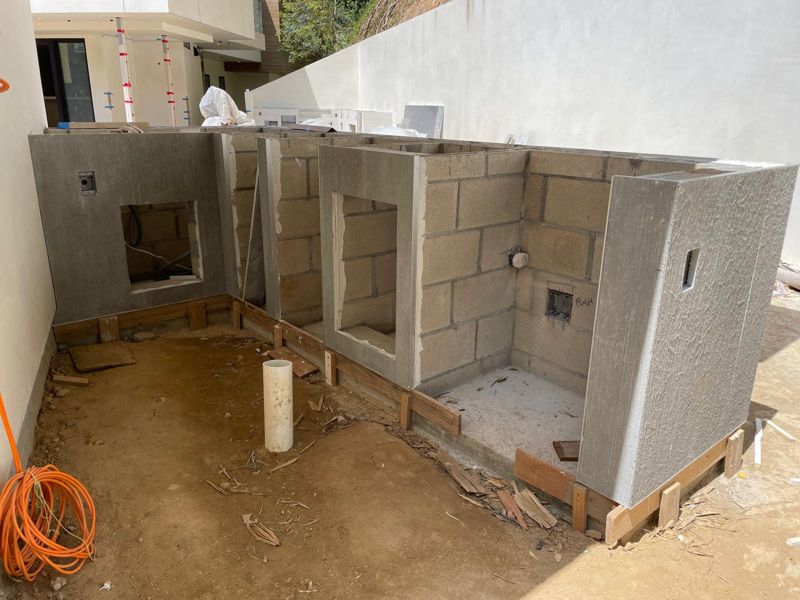 Concrete block outdoor kitchen island framed with appliance openings on a dirt pad beside a modern house