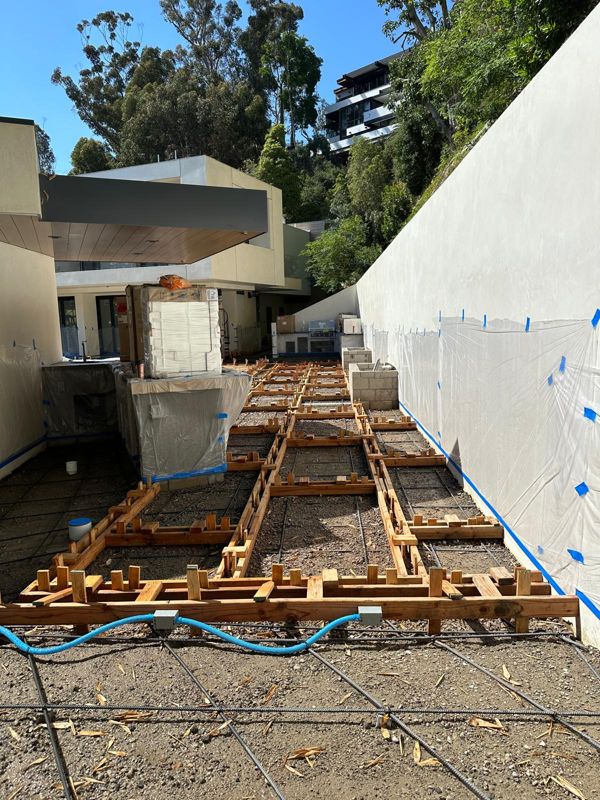 Rebar and wood forms laid out for a grid of concrete pads on gravel beside a modern house
