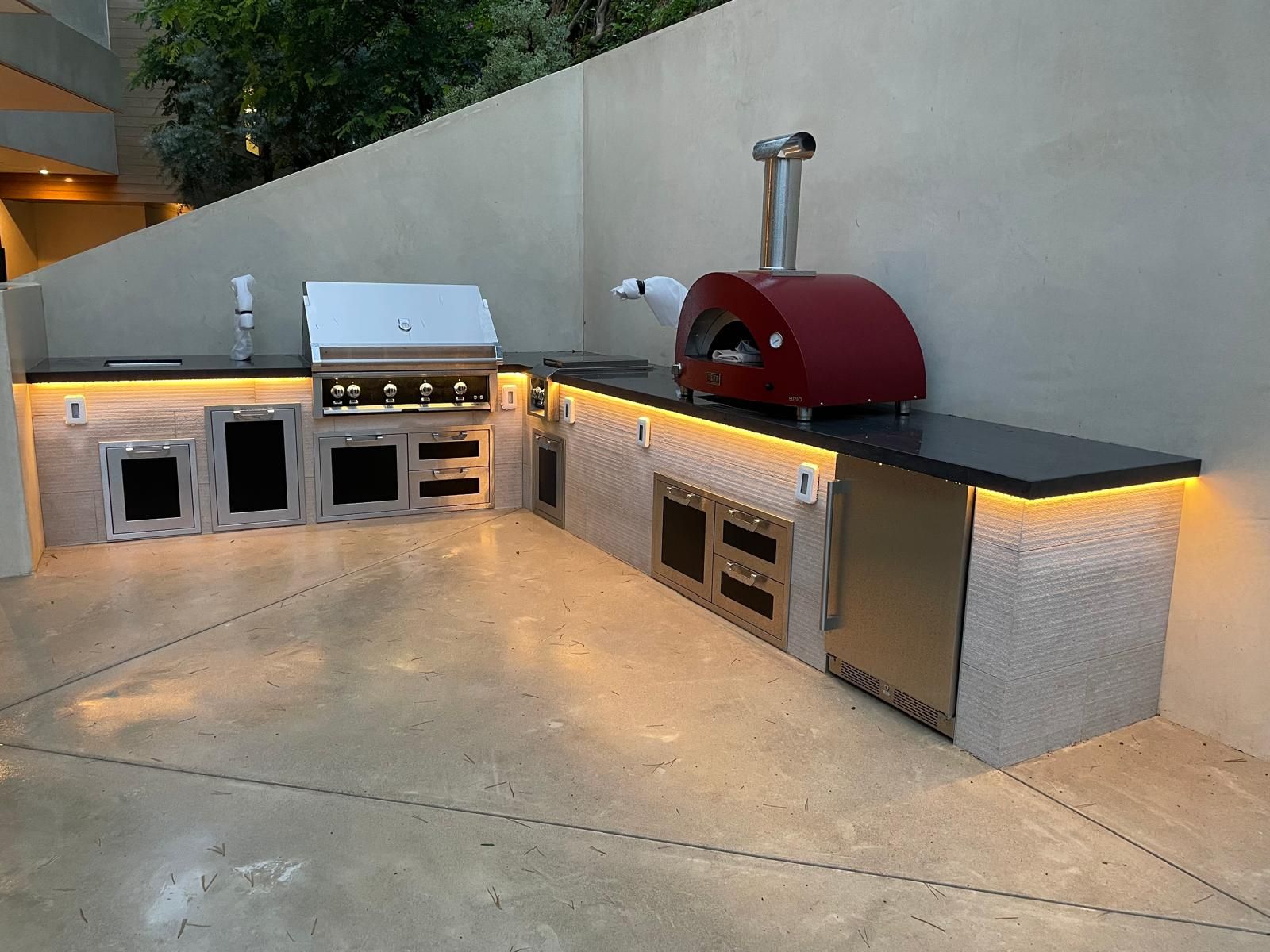 Finished L-shaped outdoor kitchen at dusk with a light stone base, dark counters, a built-in grill and a pizza oven