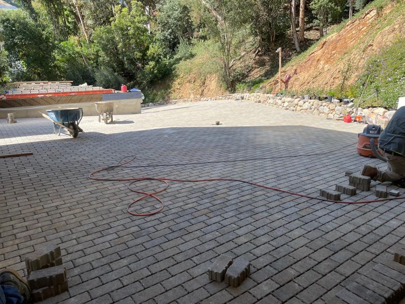 Large paver motor court mostly laid, with a boulder wall and a wooded hillside behind