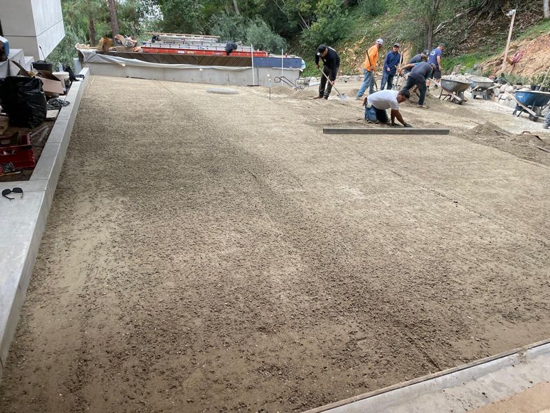 Our crew raking and screeding a sand bedding layer across a large pad beside a concrete wall