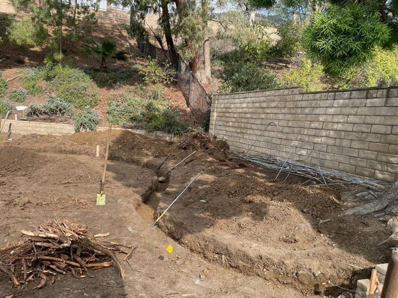 Backyard slope being graded with a curved trench cut and drip tubing coiled by the block wall