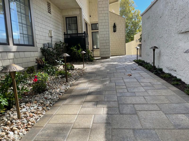 Finished paver walkway along a cream shingle house with river-rock beds, new shrubs and path lights