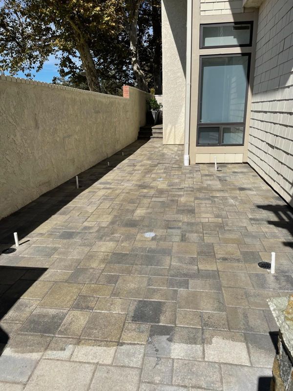 Newly laid paver patio between a stucco wall and a shingle house, with drain grates set flush