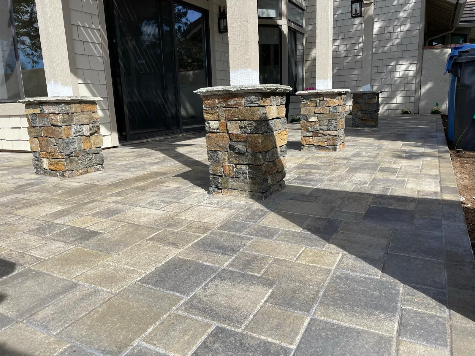 Finished paver patio with stacked-stone clad column bases under pergola posts in front of a cream shingle-sided house