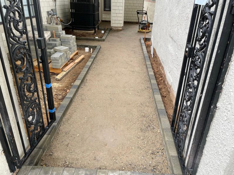 Side-yard base prep with paver borders set on both sides, seen through an iron gate