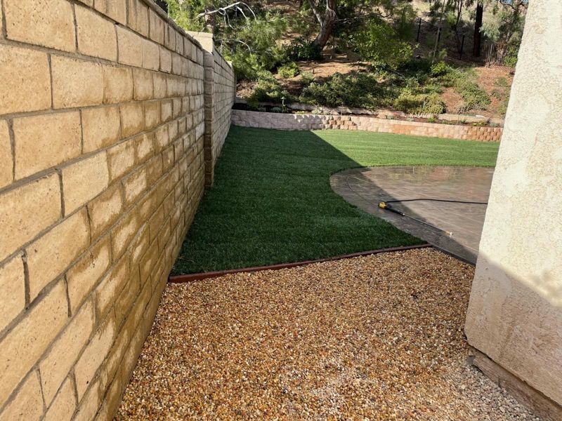 New sod along a block wall meeting a curved stamped-concrete patio and a pebble bed
