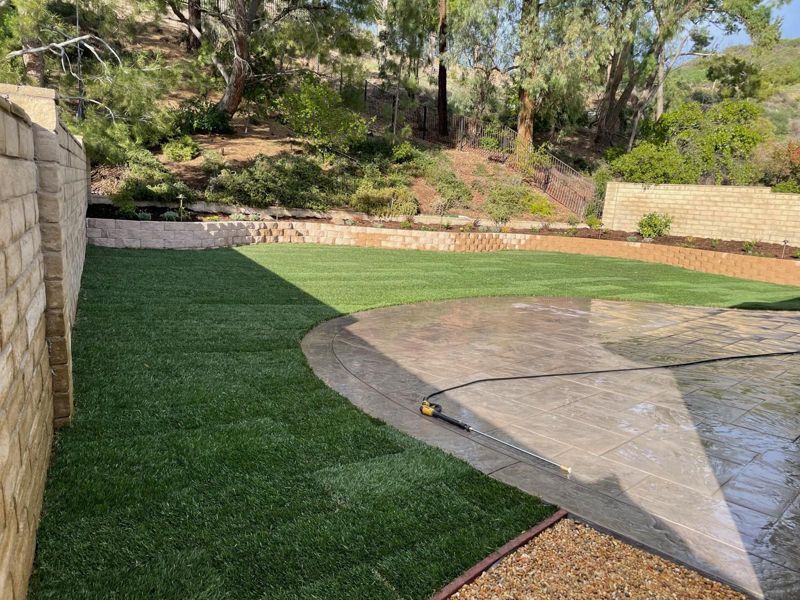 Finished backyard with fresh sod, a curved stamped patio and a planted block retaining wall
