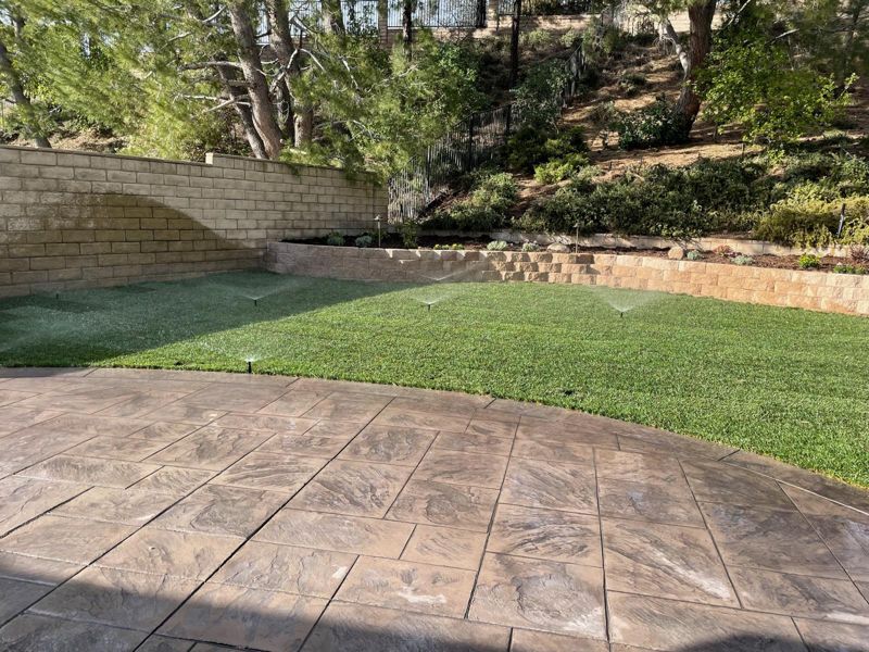 Sprinklers running on new sod beside a stamped-concrete patio and a planted retaining wall