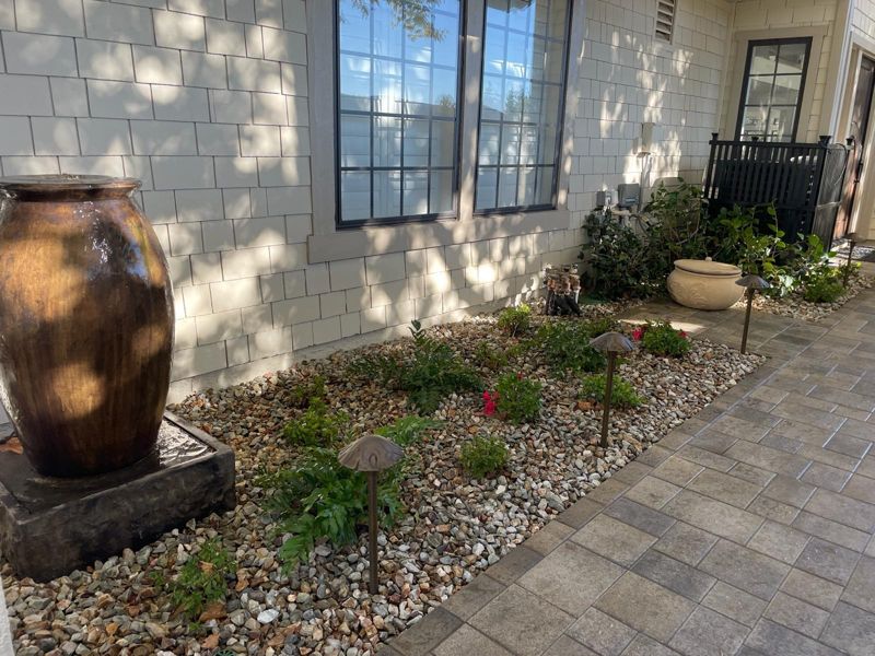 Gravel planting bed with a glazed urn fountain, small shrubs and path lights beside a paver walk