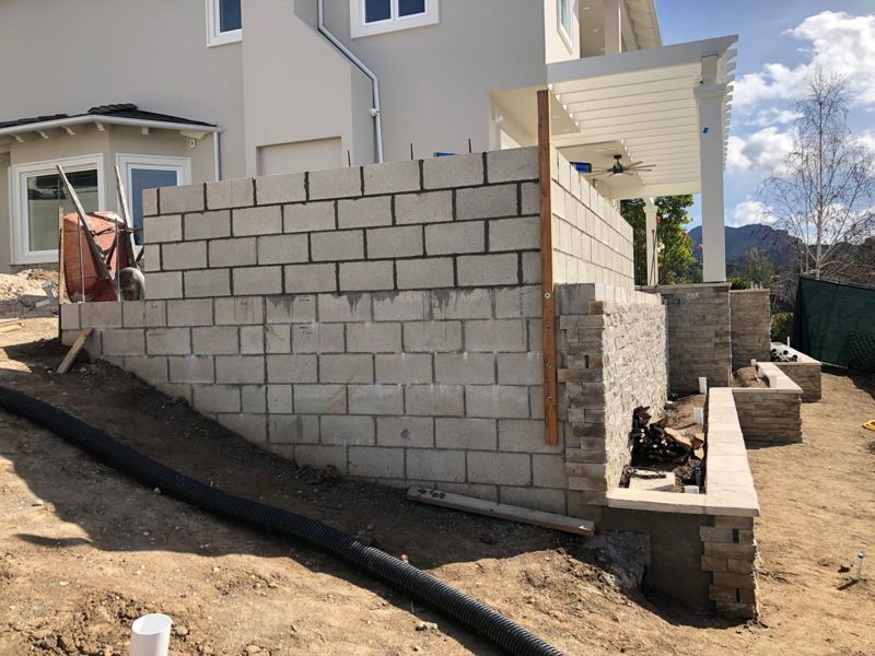Tall block wall with stacked-stone veneer partly applied and a drain pipe in the trench