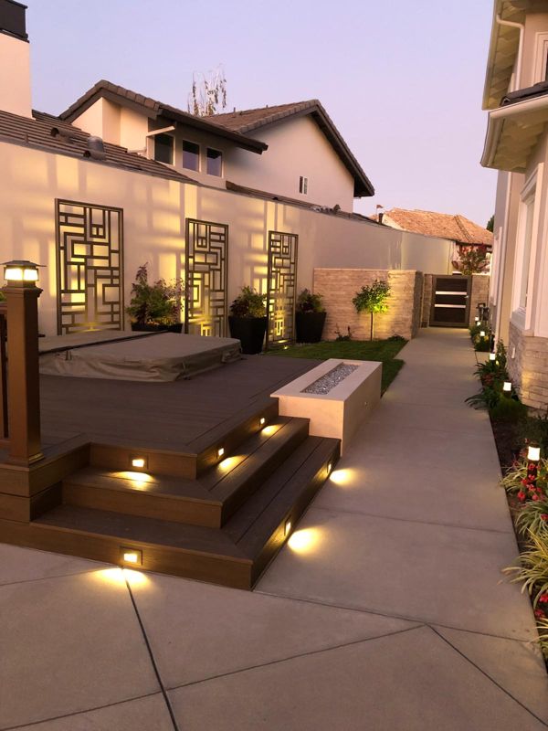 Side yard at dusk with lit deck steps, a linear fire pit, bronze wall screens and low path lights along a concrete walk