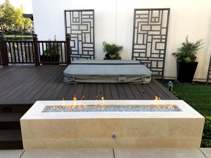 Finished linear travertine fire pit lit on a dark composite deck with black planters