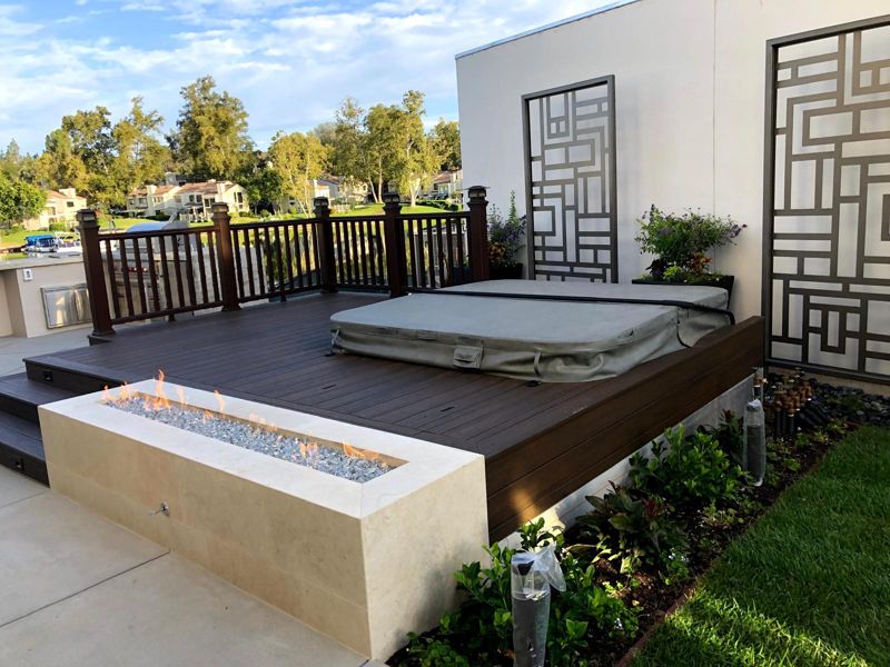 Raised composite deck with a lit linear travertine fire pit and bronze screens beside a lake
